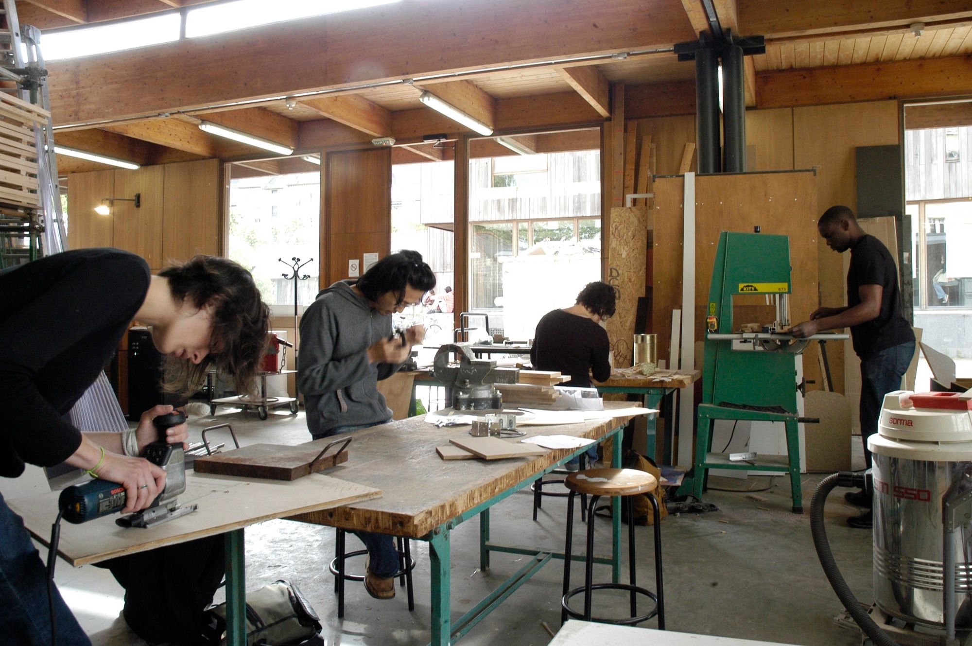 Taller de maquetas con personas cortando piezas con sierra de calar y sierra de cinta sobre mesas de trabajo